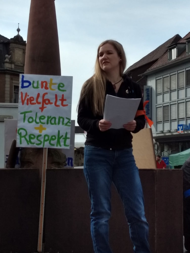Annette Linder auf dem Marktplatzbrunnen beim Vortragen eines Statements, welches sie von einem Zettel abliest. Hinter ihr steht ein Transparent mit der Aufschrift „bunte Vielfalt + Toleranz + Respekt“.