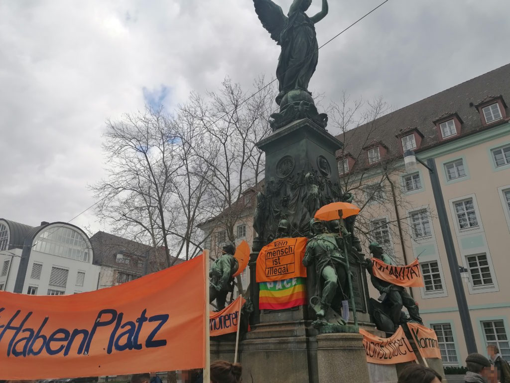 Das Siegesdenkmal auf dem Europaplatz ist mit orangefarbenen Transparenten und Regenschirmen bestückt. Auf einigen Transparenten kann man „kein mensch ist illegal“ oder „Humanität“ lesen.